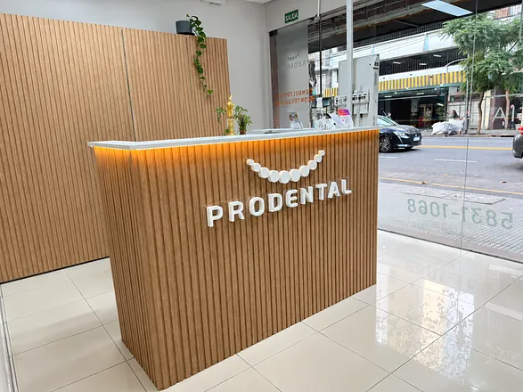 Recepción con el mostrador de listones de madera iluminado desde abajo, el logo y la palabra PRODENTAL, y la vidriera a la calle
