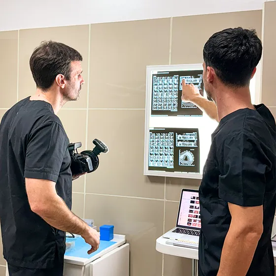 Dos profesionales de ambo negro revisando radiografías en un negatoscopio, uno de ellos con una cámara de fotos