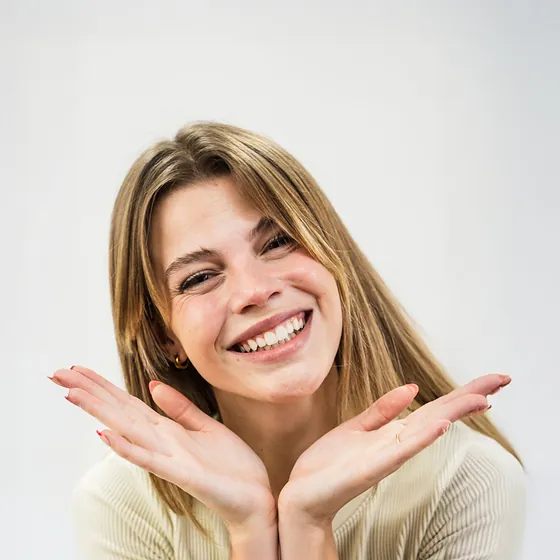 Retrato de estudio de una mujer de pelo largo y remera clara, sonriendo, con las dos manos abiertas debajo del mentón