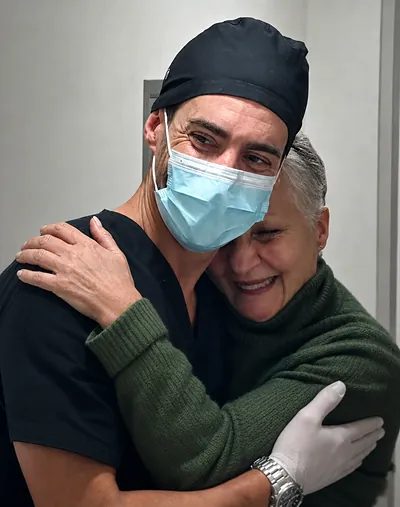 Un profesional de Prodental con barbijo y gorro abraza a una paciente de pelo canoso y suéter verde, que sonríe con los ojos cerrados
