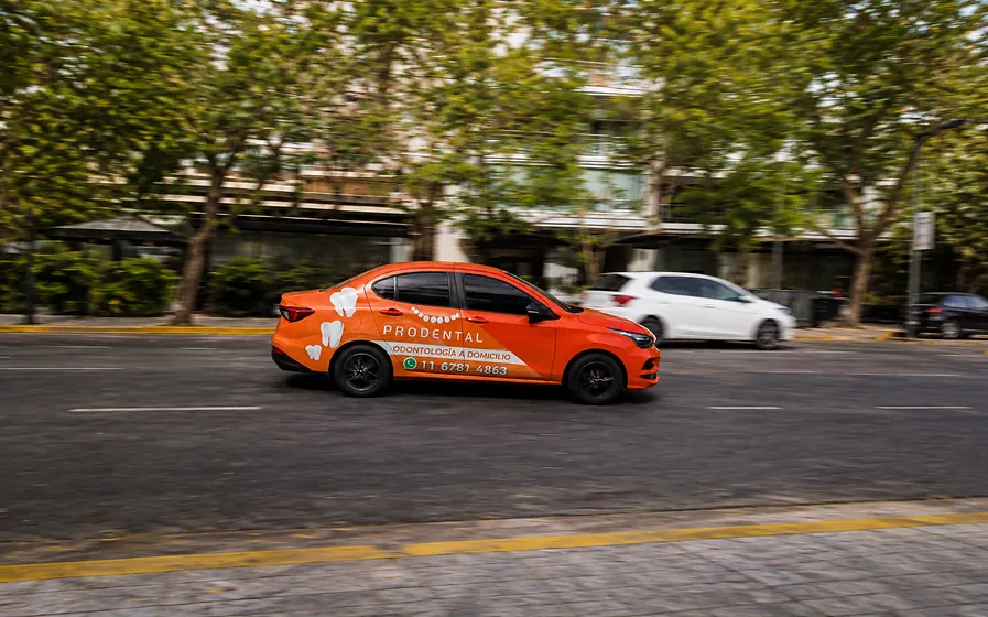 Un auto de Prodental rotulado en naranja, con la leyenda Odontología a domicilio, circulando por una avenida arbolada