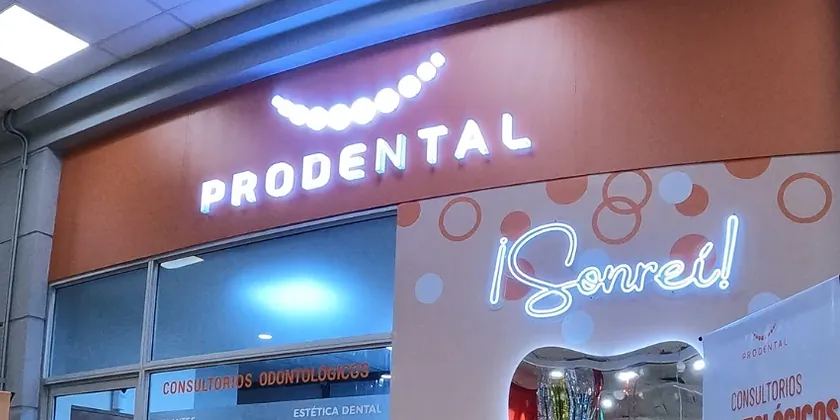 Frente de una sucursal de Prodental con el cartel PRODENTAL iluminado sobre la fachada naranja y el letrero ¡Sonreí! en neón